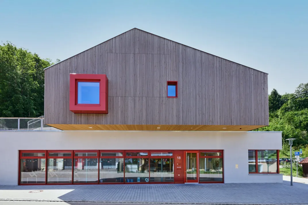 Straßenansicht mit Holzfassade, rotem Fenstererker und verglastem Erdgeschoss